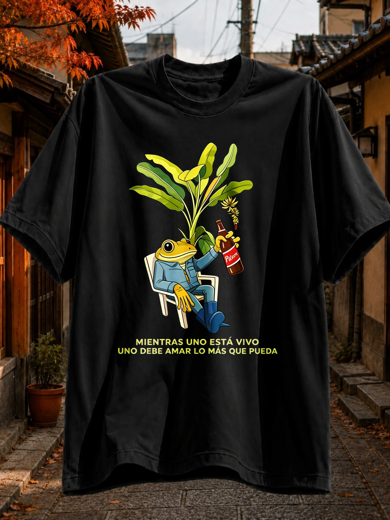 Tant qu'on est vivant, il faut aimer plus que tout autre chose - T-shirt noir, grenouille de dessin animé détendue tenant une bouteille de Pilsen à côté d'une plante verte luxuriante.