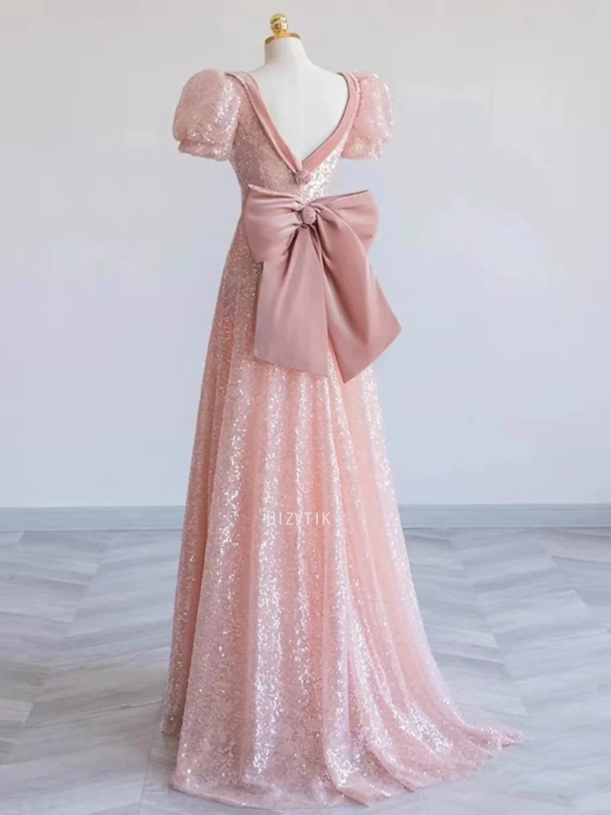 ชุดราตรีสีชมพู แขนพองสั้น คอวี ประดับเลื่อม แต่งโบว์ พร้อมระบายชายกระโปรงยาวลากพื้น สำหรับงานพรอมหรืองานเลี้ยงอย่างเป็นทางการ