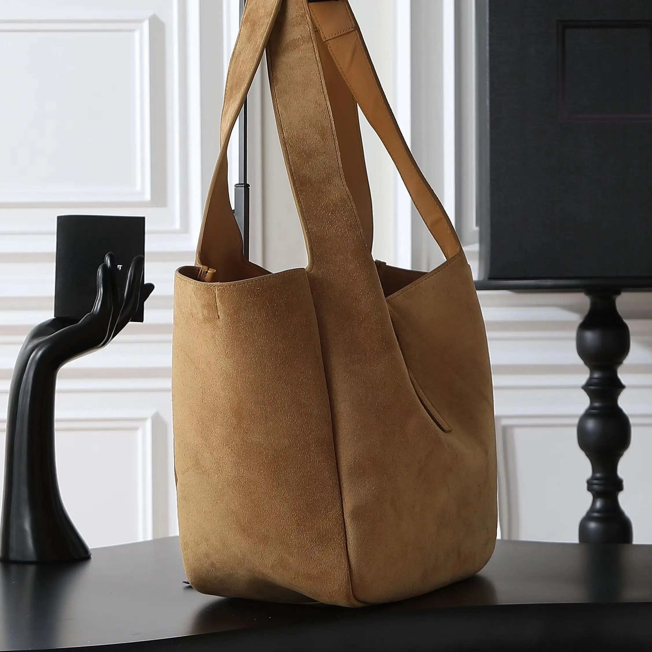 

Tan suede open-top tote bag with a wide single shoulder strap, minimalist central buckle, and soft draped silhouette.