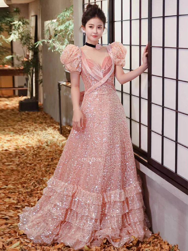 ชุดราตรีสีชมพู แขนพองสั้น คอวี ประดับเลื่อม แต่งโบว์ พร้อมระบายชายกระโปรงยาวลากพื้น สำหรับงานพรอมหรืองานเลี้ยงอย่างเป็นทางการ