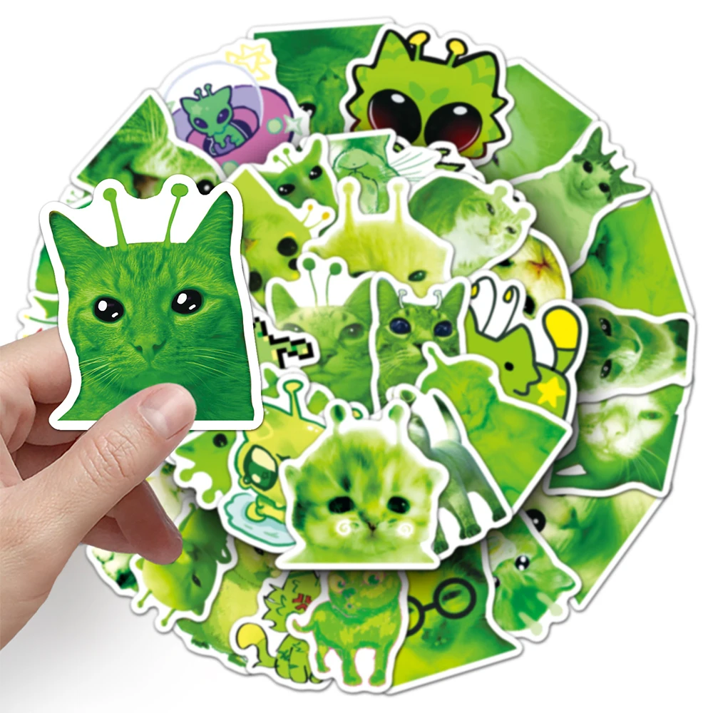 Hand holding a green cat sticker with alien antennae in front of an assortment of cat stickers