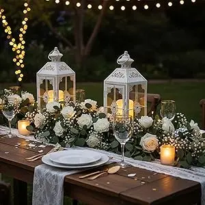 Elegant outdoor table setting with white lanterns, floral arrangements, and candles creating ambient lighting at dusk. Table decorated with greenery and place settings.