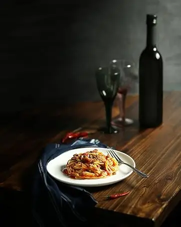 Plate of pasta dish on wooden table with wine bottle, glasses, and moody lighting, creating a restaurant-like ambiance.