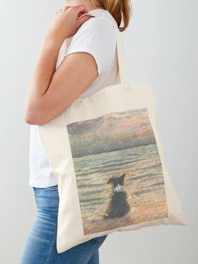 A Border collie dog gazes out over the ocean watching the sun come up, greeting the morning Tote Bag