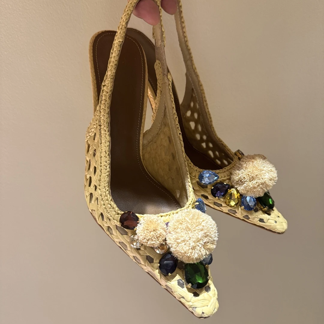 Tacones altos de mujer con punta abierta y tejido de rafia, adornados con gemas y pompones en los tacones de aguja delgados.