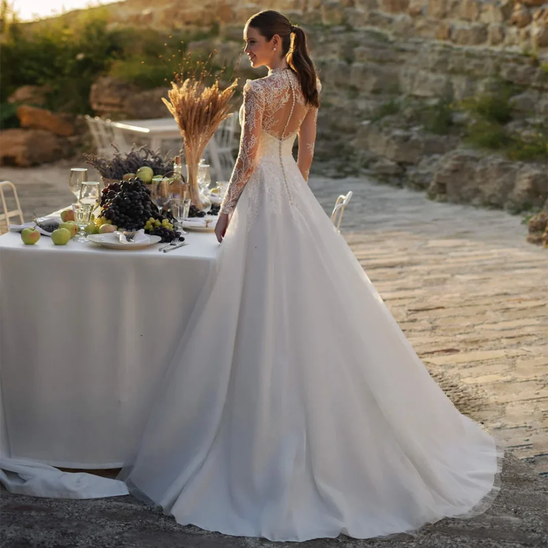 Elegancka suknia ślubna szyta na miarę, z wysokim dekoltem, długimi rękawami, iluzoryczną koronką, aplikacjami, tiulową spódnicą w kształcie litery A, trenem zamiatającym.