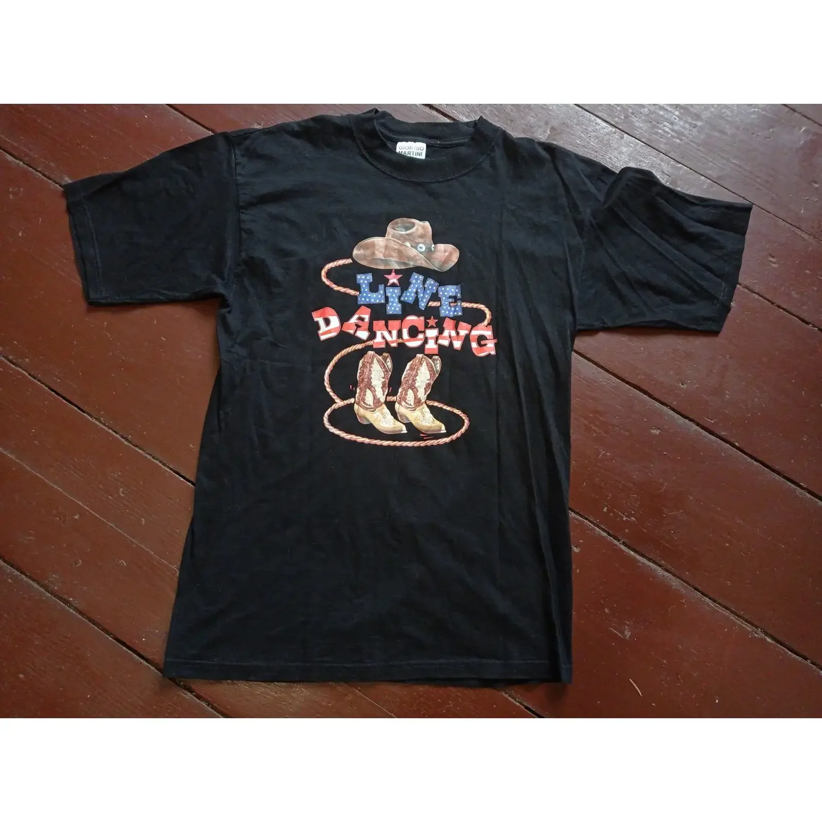 May include Black t shirt featuring a graphic of cowboy hat boots and the text Line Dancing in red white blue vintage Washed
