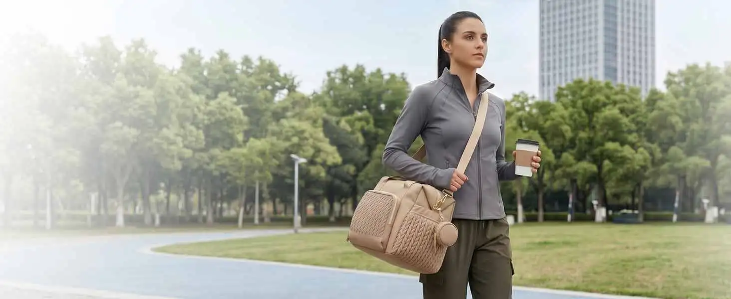 Man in business casual attire (gray blazer, tan pants) wearing brown shoulder bag, standing outdoors in park setting with trees and water visible in background.