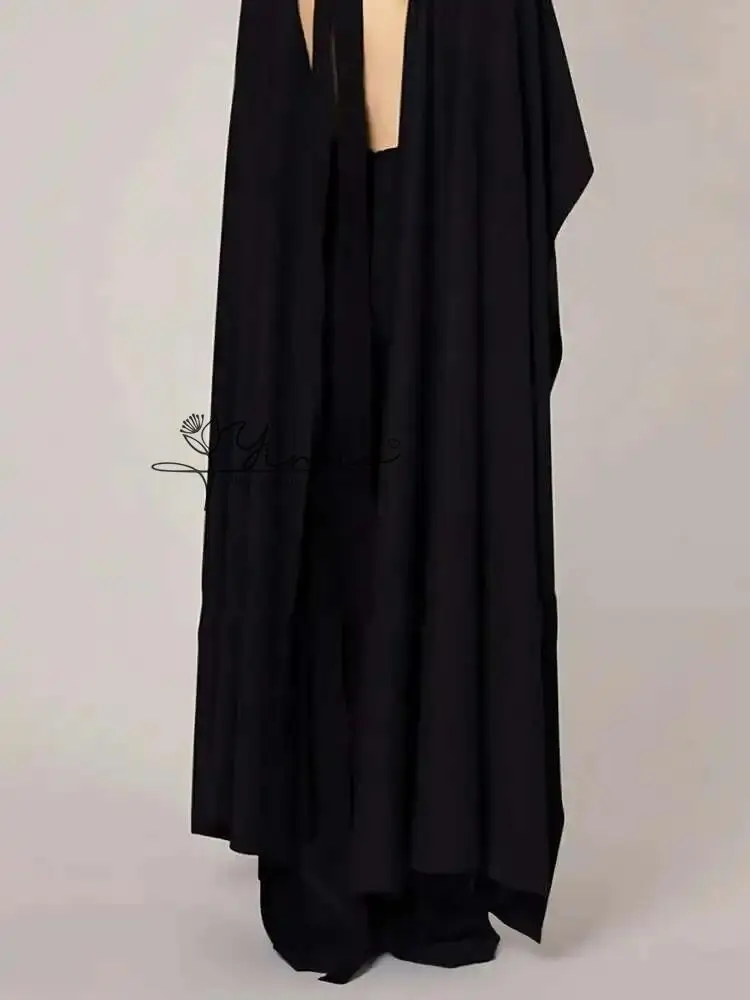Czarny kombinezon na zamówienie z peleryną, golfem, szerokimi nogawkami, zwiewnymi rękawami, eleganckim kreponem i bocznymi kieszeniami.
