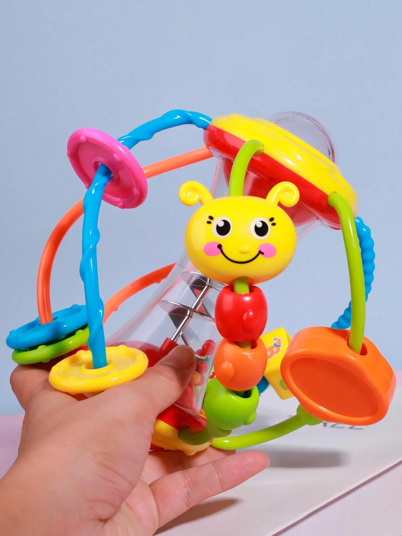 Randomly colored. A rattle activity ball toy designed for infants aged 12 months, featuring swaying arms and rotating rattles.