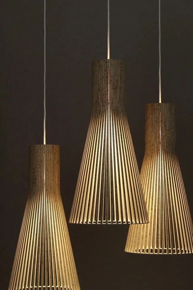 Three identical pendant lights hang against a dark background. The lights have a cylindrical, slatted design and emit a warm glow, creating a pattern of light and shadow.