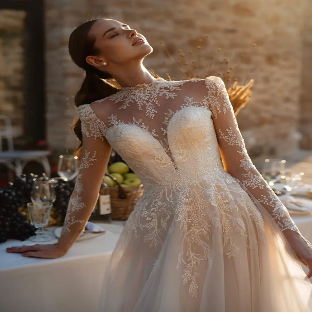 Elegancka suknia ślubna szyta na miarę, z wysokim dekoltem, długimi rękawami, iluzoryczną koronką, aplikacjami, tiulową spódnicą w kształcie litery A, trenem zamiatającym.