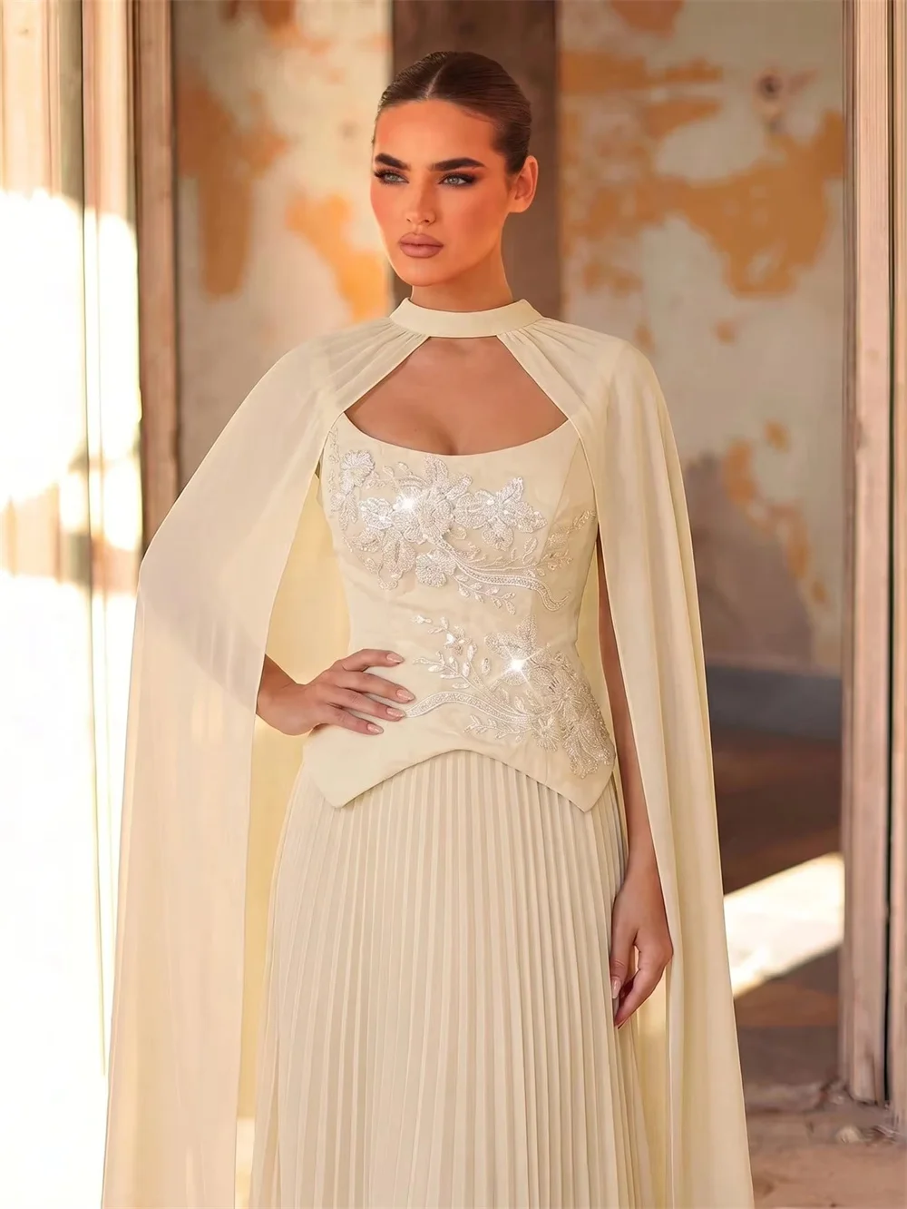 

Cutout square neck gown with sheer cape, fitted waist and flowing pleated fabric, floor-length elegant skirt. Minimalist luxury