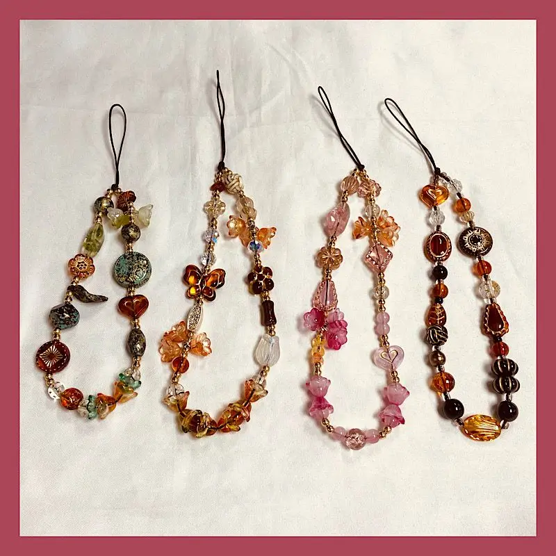Quatre lanières de charme de téléphone en perles faites à la main, présentant des perles de verre et de résine aux tons ambre, corail, rose et bronze, avec des motifs floraux et en forme de cœur finement détaillés, destinées à être utilisées comme ornements d’accrochage au poignet.