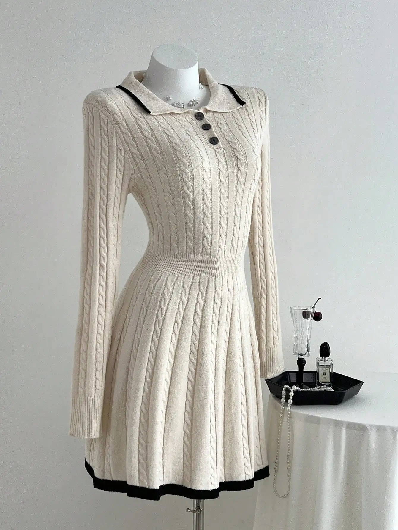 Damska kremowa sukienka swetrowa z długim rękawem, z kołnierzykiem polo, dzianiną w warkocze i czarnymi wykończeniami.