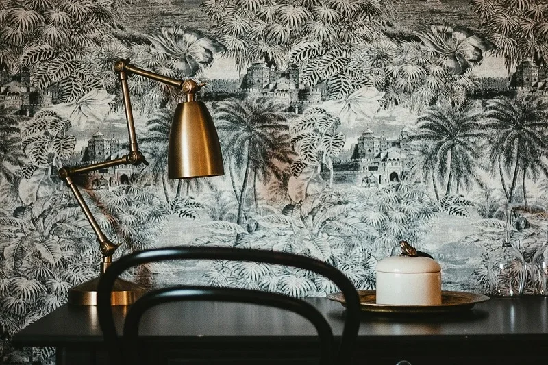 A brass desk lamp illuminates a small white jar on a gold tray atop a dark desk. Behind, a black chair faces a black-and-white tropical toile wallpaper featuring elephants and architecture.
