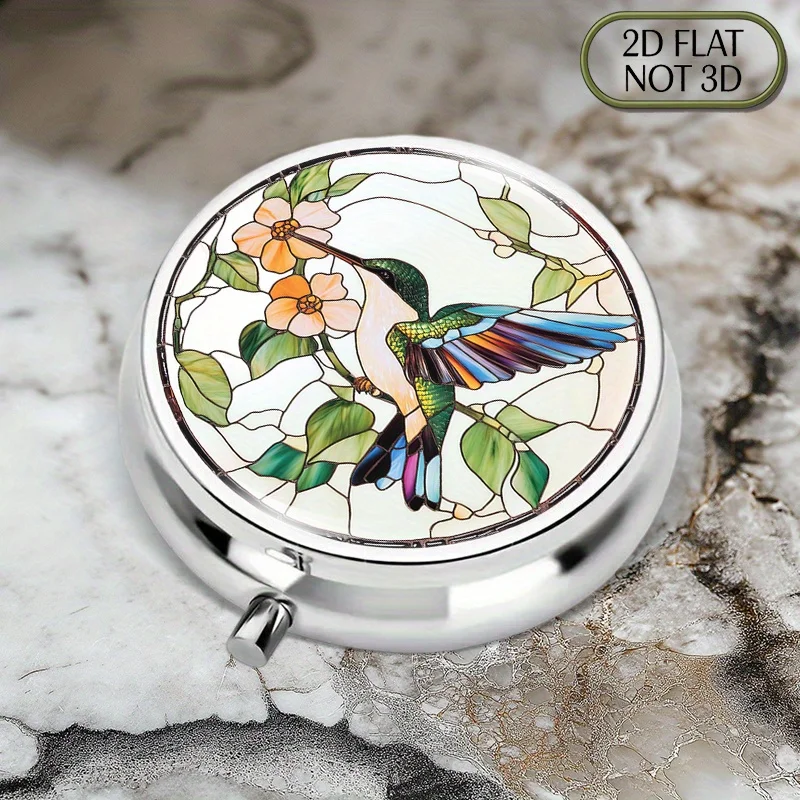 A flat, portable, round metal pill box with a painted glass hummingbird and flower pattern, suitable for organizing medication and small essentials. It is a perfect choice for storing vitamins, small accessories, or as a thoughtful summer gift for loved ones